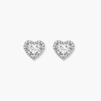 Close-up of heart-shaped moissanite stud earrings in sterling silver halo setting on cream background.