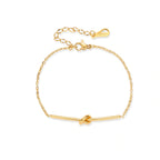 Delicate gold chain bracelet with minimalist bar and centered knot detail on cream background