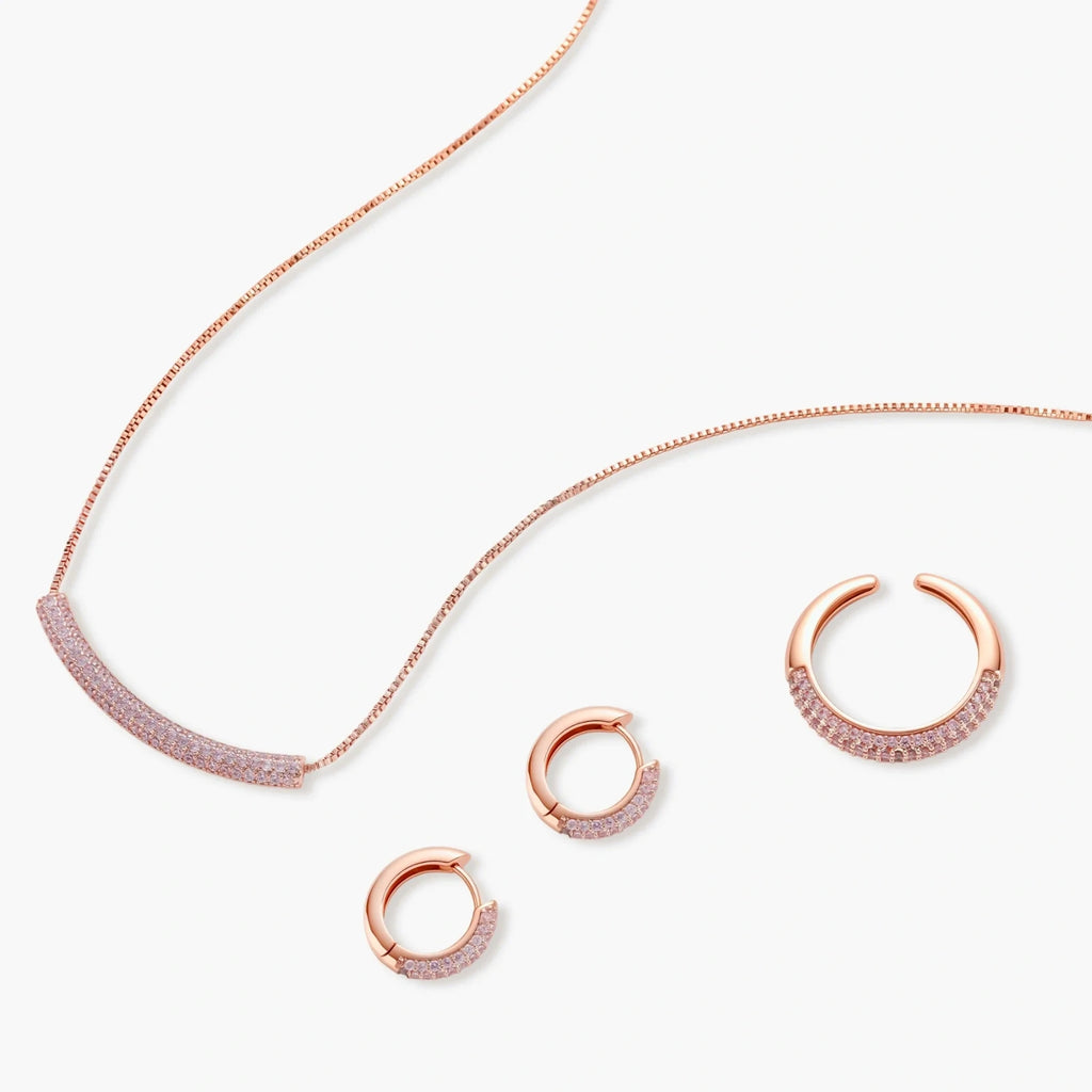 Rose gold pavé zirconia bar necklace, hoop earrings, and open ring set with pink stones on cream background.